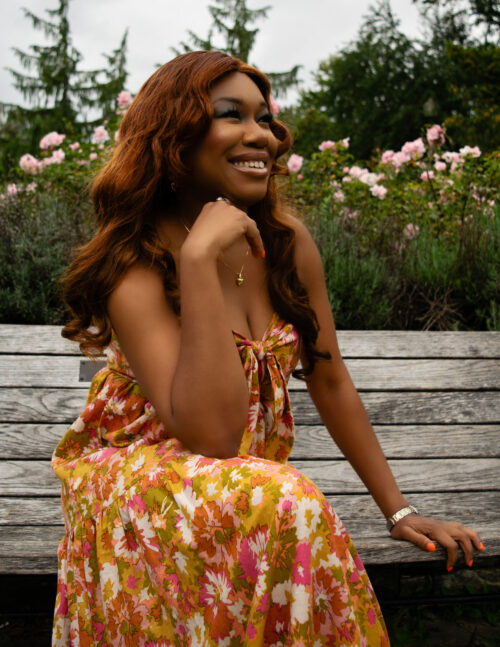 A woman wearing a floral dress sitting on a bench, smiling sideways.